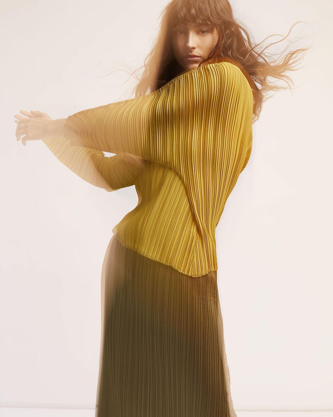Woman in pleated gold blouse and long skirt poses in a studio, arms raised, exuding editorial fashion elegance and style.