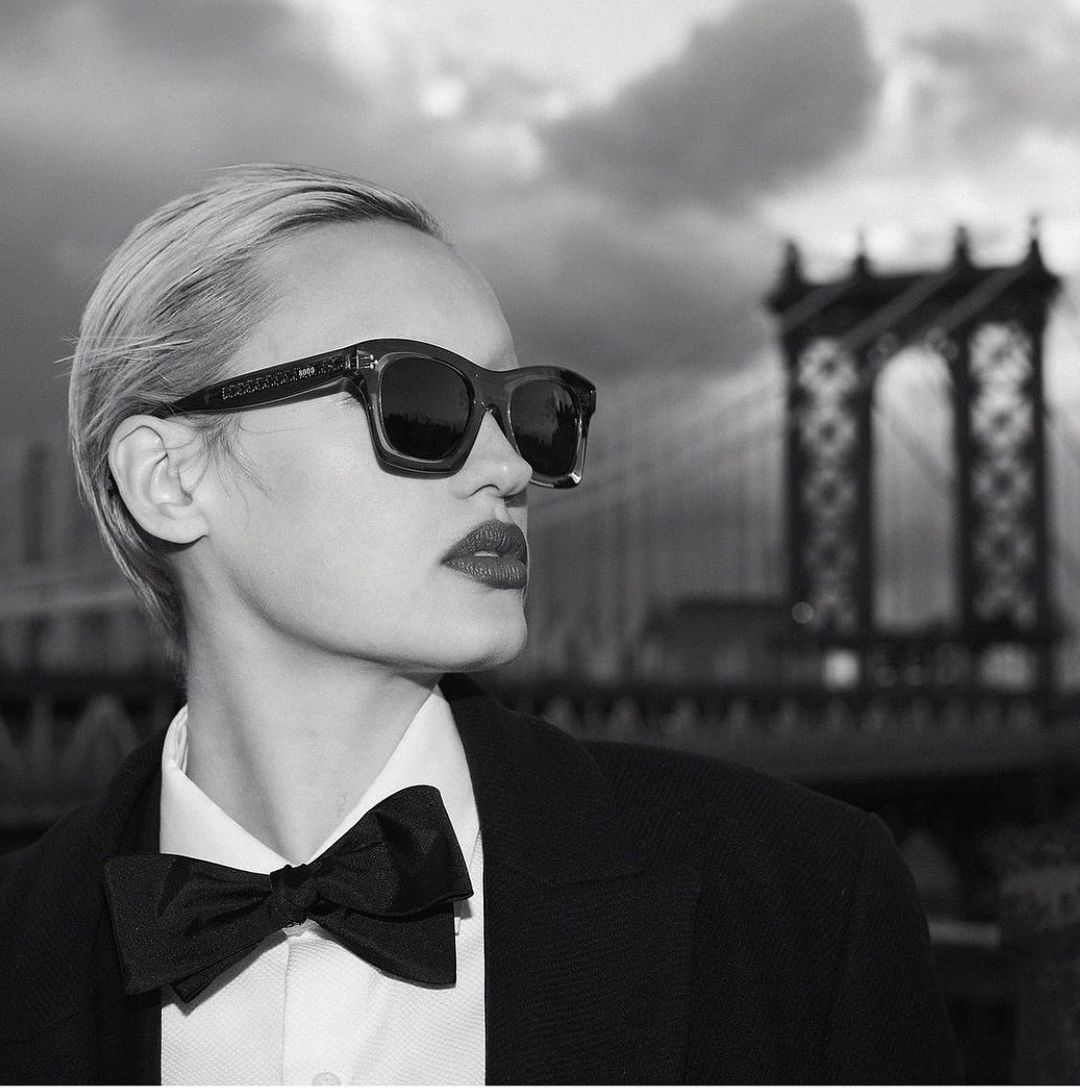 Monochrome portrait of a woman wearing dark sunglasses, a white collared shirt, black bow tie, and dark jacket with a bridge in background.