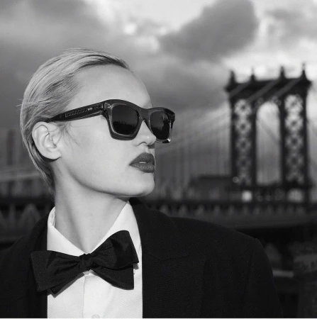 Monochrome portrait of a woman wearing dark sunglasses, a white collared shirt, black bow tie, and dark jacket with a bridge in background.