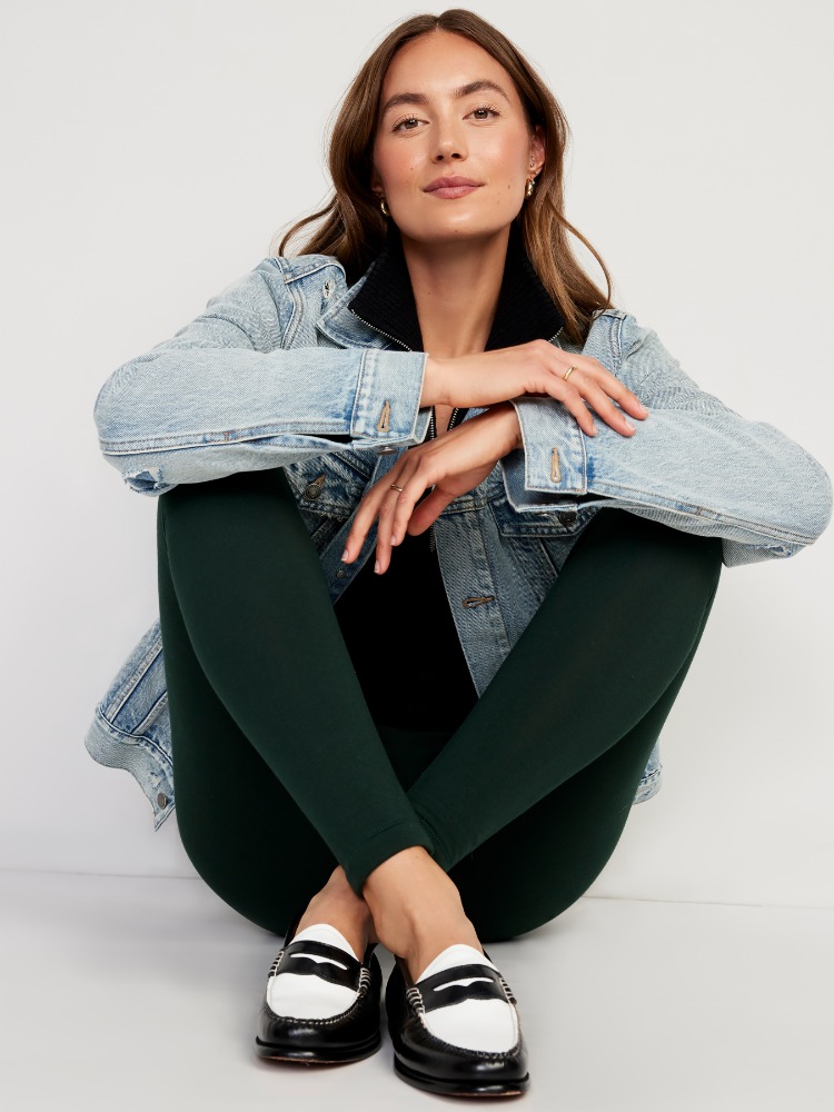 Attractive woman poses indoors wearing a light wash denim jacket, dark leggings, and black and white penny loafers on a white floor.