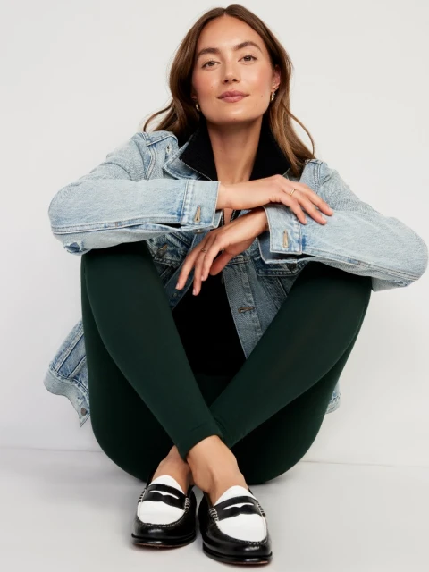 Attractive woman poses indoors wearing a light wash denim jacket, dark leggings, and black and white penny loafers on a white floor.