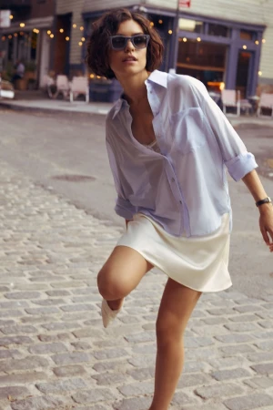 Woman wearing sunglasses, light blue button-up shirt, and white skirt on a cobblestone street by Caitlin Mitchell