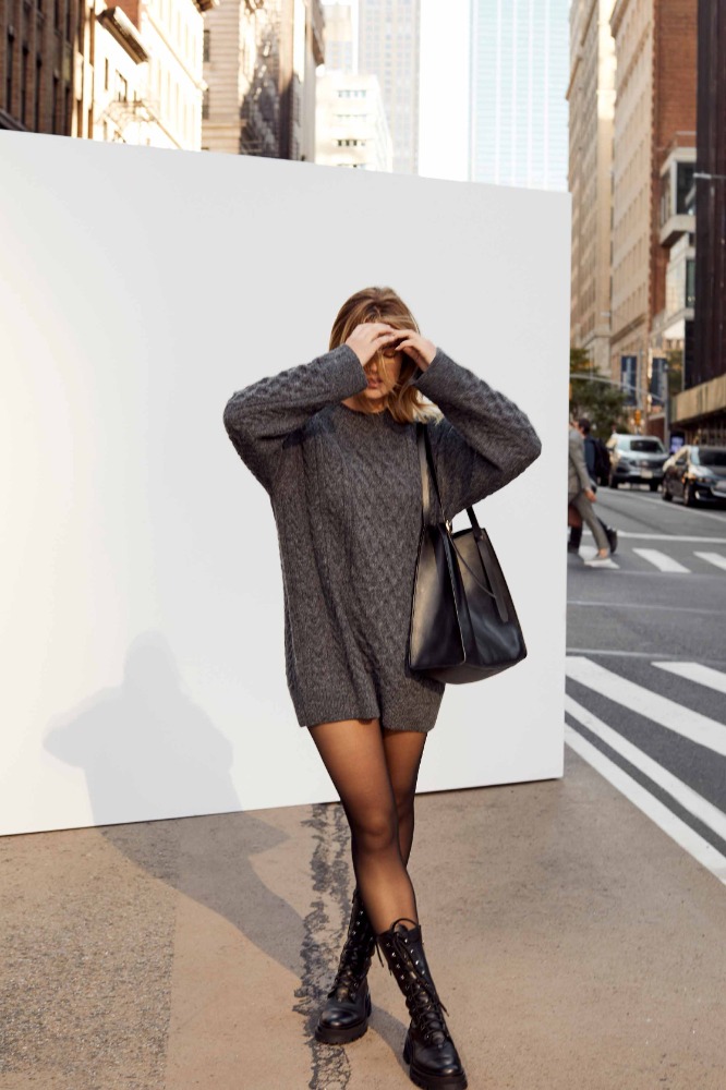 Woman in grey sweater dress, black tights, and combat boots stands against a white wall in an urban setting with high-rise buildings.