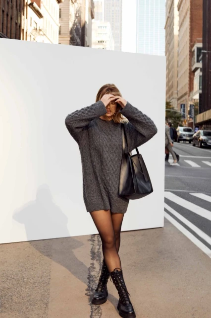 Woman in grey sweater dress, black tights, and combat boots stands against a white wall in an urban setting with high-rise buildings.