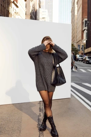 Woman in grey sweater dress, black tights, and combat boots stands against a white wall in an urban setting with high-rise buildings.