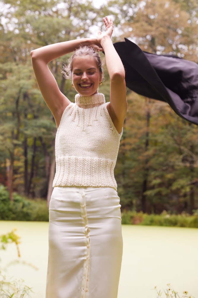 Smiling woman with arms raised outdoors, wearing a sleeveless cream-colored knit top and cream skirt near greenery and trees in natural light.