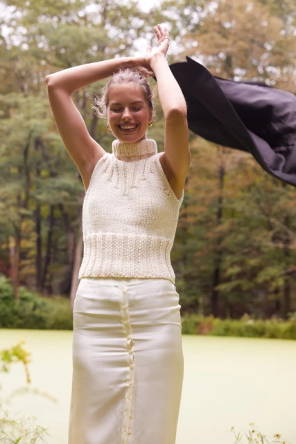 Smiling woman with arms raised outdoors, wearing a sleeveless cream-colored knit top and cream skirt near greenery and trees in natural light.