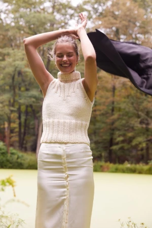 Smiling woman with arms raised outdoors, wearing a sleeveless cream-colored knit top and cream skirt near greenery and trees in natural light.