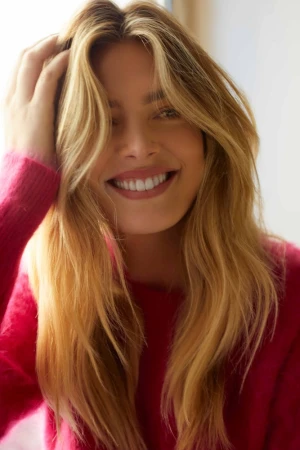 Close-up portrait of a smiling adult woman with long blonde hair, wearing a pink sweater, with her hand in her hair, and a blurred background.