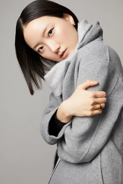 Portrait of an Asian woman wearing a grey wool coat, hugging herself, looking at the camera on a neutral grey background.