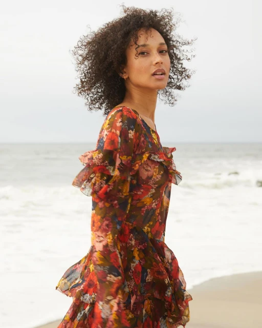 A woman with curly hair wears a flowing floral dress on a beach with waves and cloudy skies, creating a serene fashion editorial image.