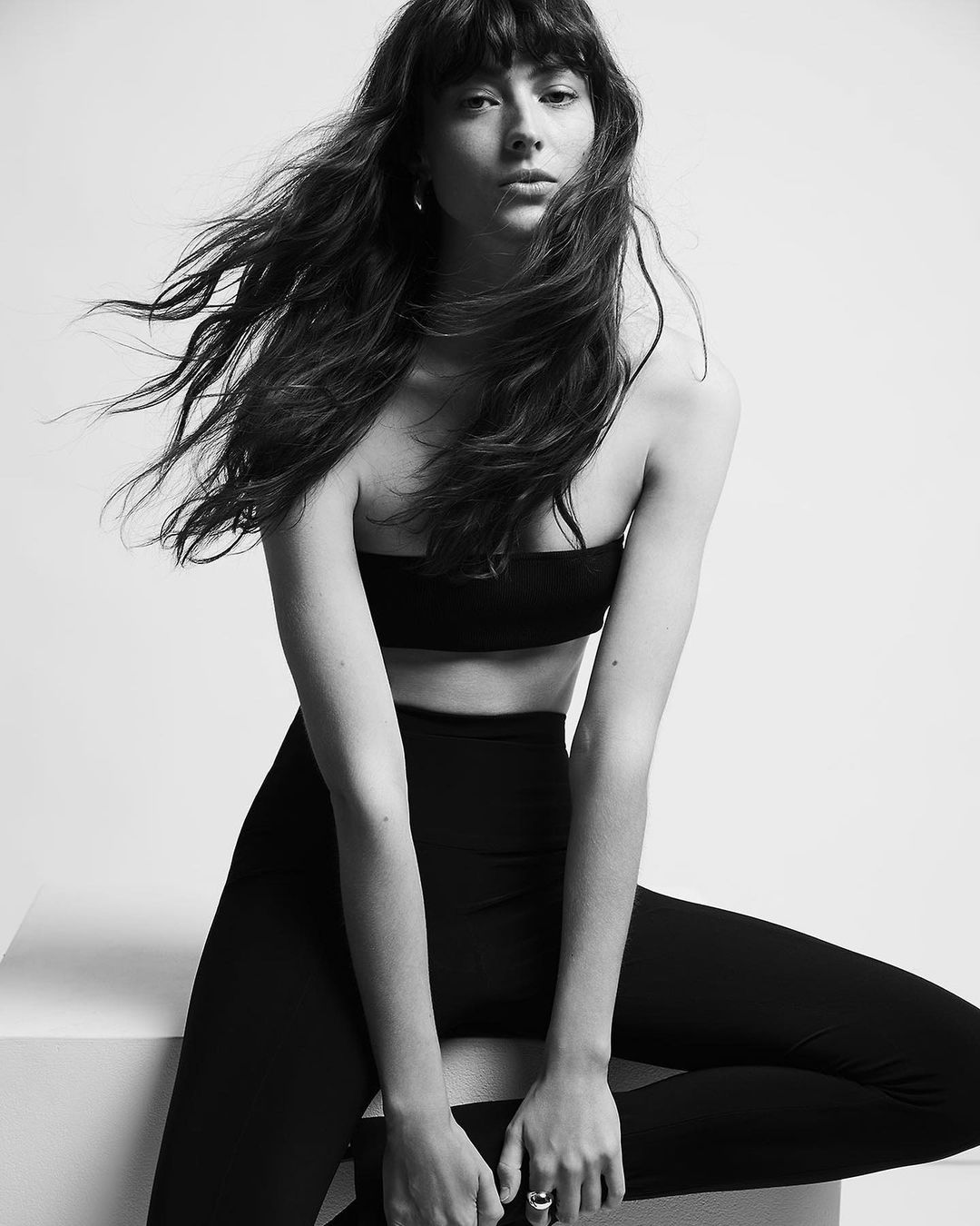 Black-and-white portrait of a brunette woman with bangs wearing a top and leggings, sitting on a white box in a photo studio.