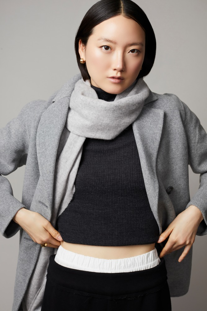 Woman in a grey coat and scarf poses in a studio. She is wearing a black top and is touching the waistband of her bottoms.