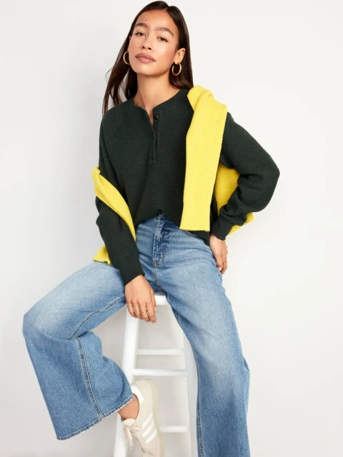 A young woman poses indoors wearing a dark-green henley shirt, yellow sweater draped over her shoulders, wide-leg jeans, and sneakers.