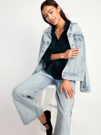 Woman wearing light wash denim jacket and matching jeans seated on a white stool against a plain background