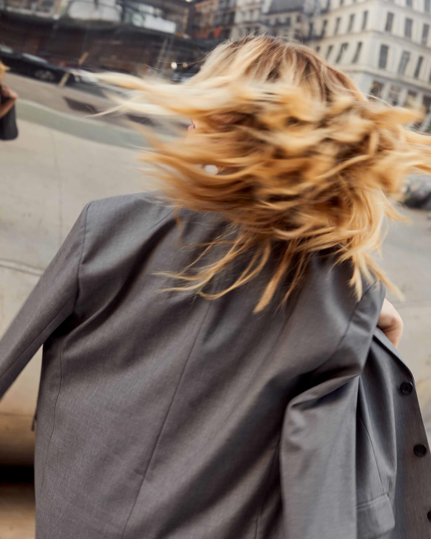 Blond hair of a woman with gray blazer blowing in the wind with blurred urban background evokes dynamism and movement.
