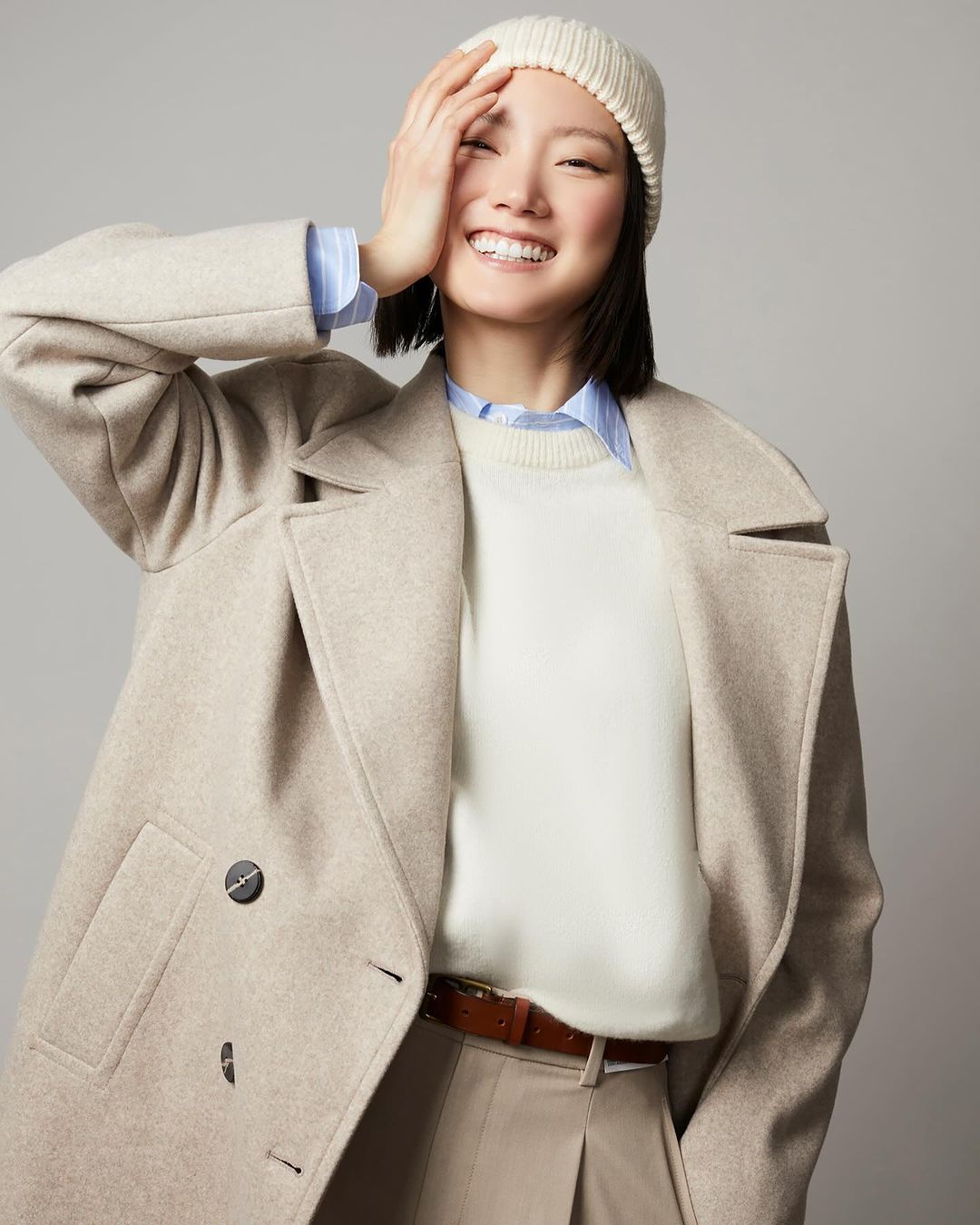 Smiling woman in beige coat, cream sweater, and knit beanie against a grey seamless studio background.