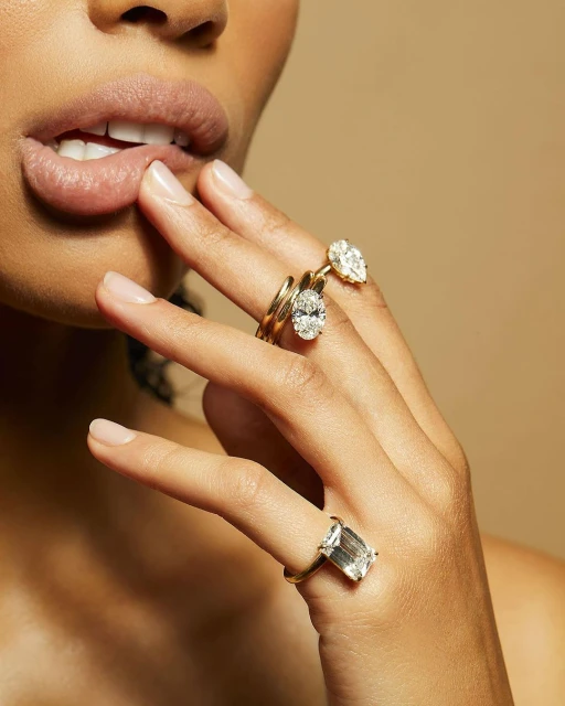 Close-up of a woman's hand adorned with three golden rings featuring diamonds. The shot is taken against a neutral backdrop.