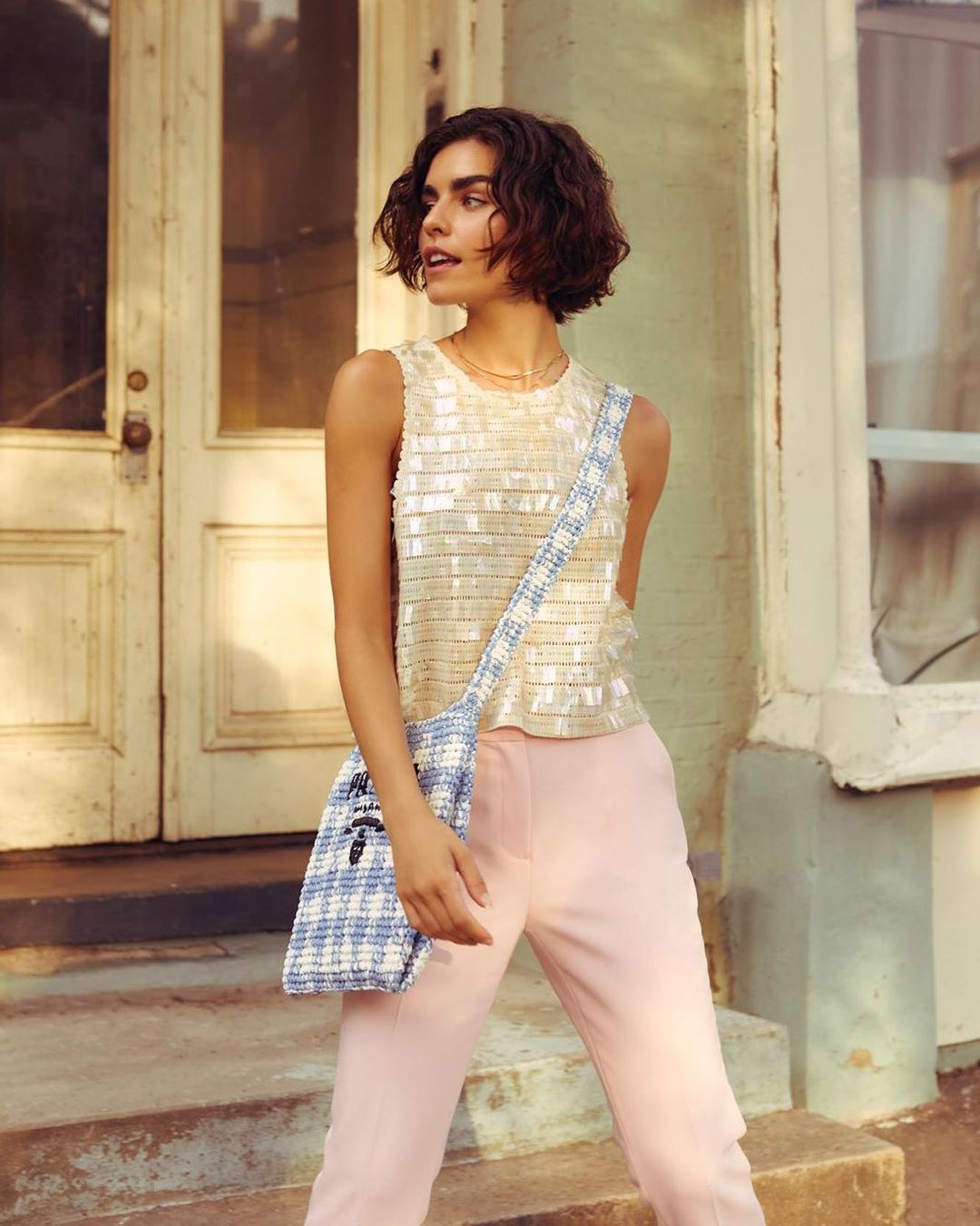 Woman in shimmering top and pink pants poses on steps in front of an aged doorway, wearing a patterned blue and white shoulder bag.