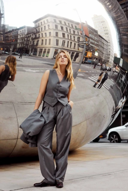 A woman in a gray vest and slacks poses on a city street with buildings and a metallic bean-shaped sculpture in the background.