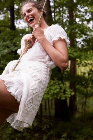 Woman on a rope swing laughing in an outdoor forest setting, wearing a white dress and knitted cardigan. Joyful and carefree.
