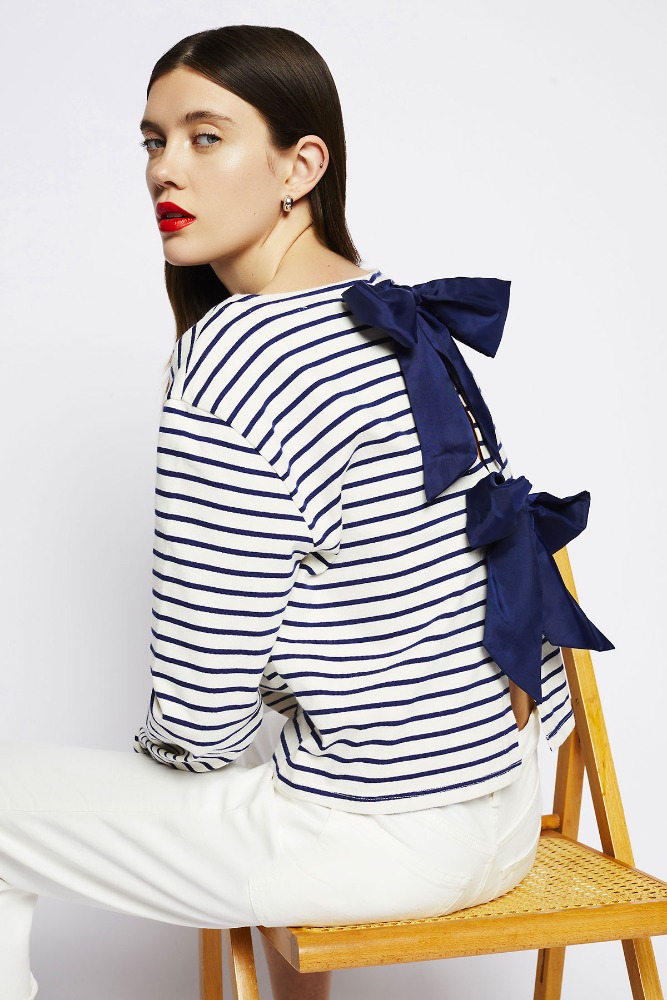 Fashion model in striped long-sleeve shirt with bow detail, sitting on a rattan chair against a white backdrop in a studio setting.