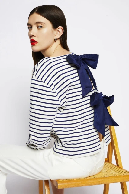Fashion model in striped long-sleeve shirt with bow detail, sitting on a rattan chair against a white backdrop in a studio setting.