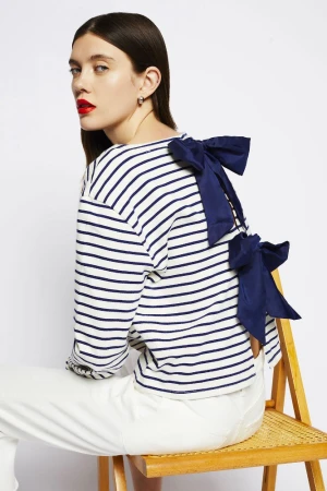 Fashion model in striped long-sleeve shirt with bow detail, sitting on a rattan chair against a white backdrop in a studio setting.