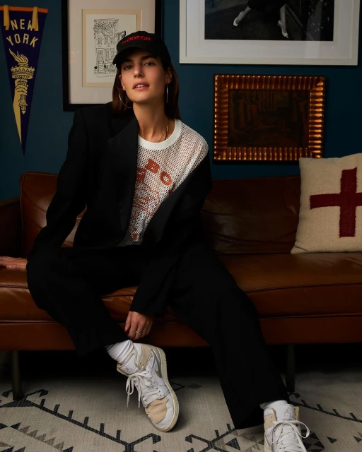 A stylish woman in a black blazer and pants sits on a brown leather couch in a cozy, indoor setting, wearing a baseball cap and sneakers.