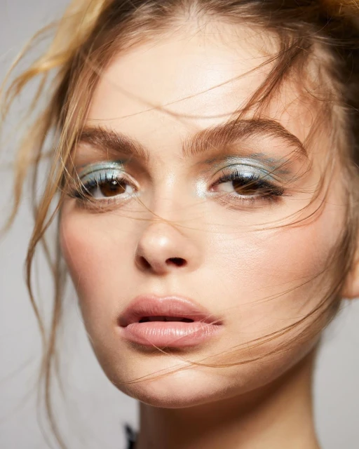 Close-up portrait of a young adult female with fair skin, light brown hair, wearing light blue eyeshadow and lip gloss, against a grey backdrop.