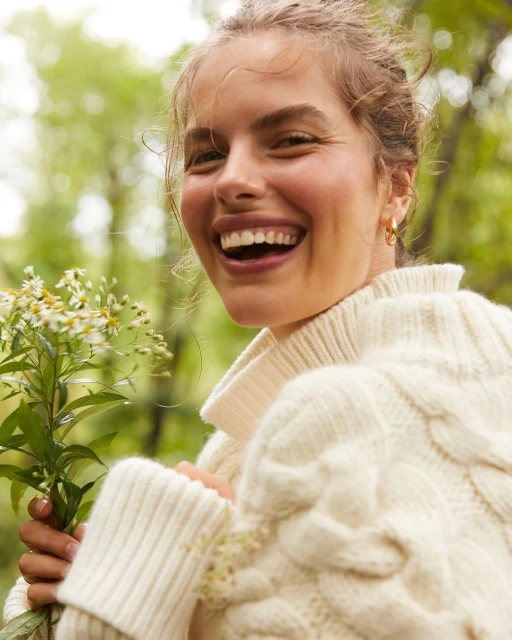 Smiling woman in a cream cable knit sweater holding a bouquet of white flowers in a green forest setting on a sunny day.