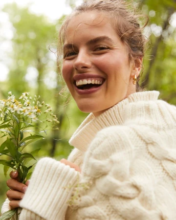 Smiling woman in a cream cable knit sweater holding a bouquet of white flowers in a green forest setting on a sunny day.