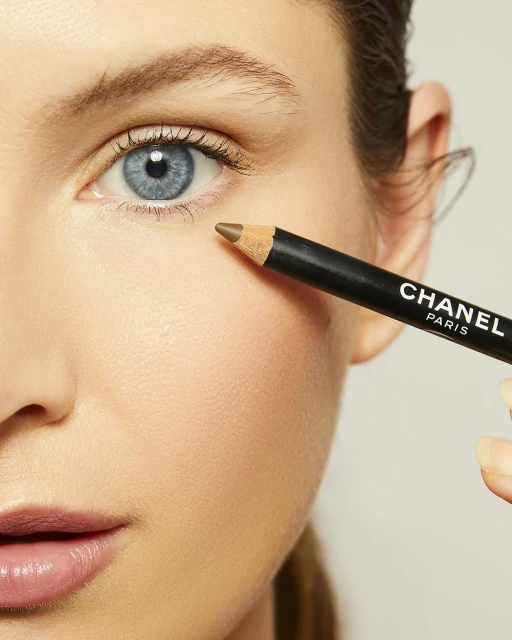 Close-up of a fair-skinned woman with blue eyes applying brown eyeliner. Cosmetic product by Chanel Paris is held by a manicured hand.