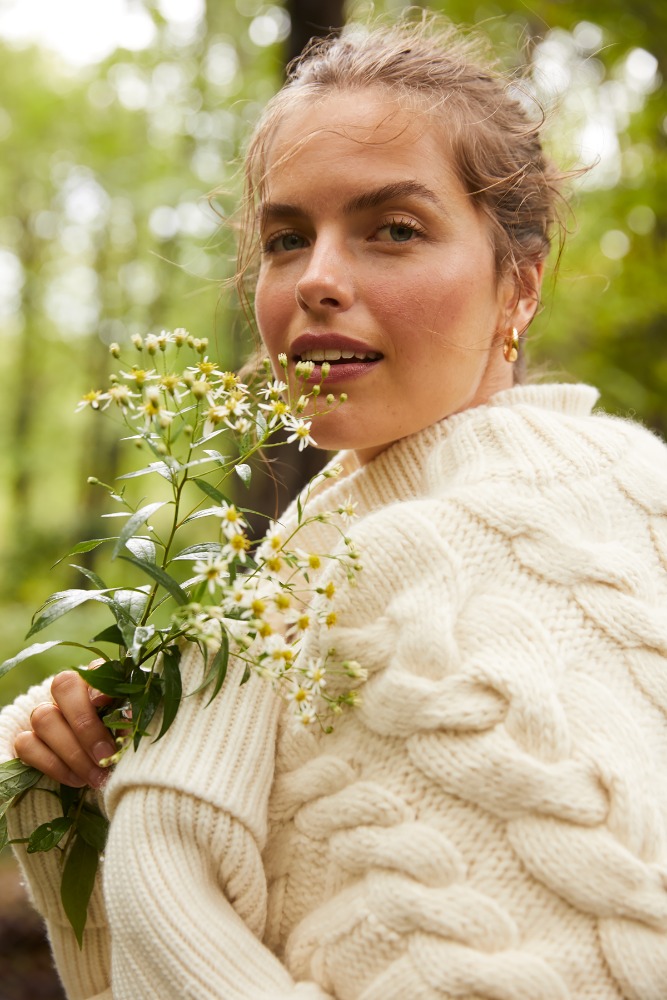 Portrait of a fair-skinned woman in a cable-knit sweater, holding a delicate sprig of small white flowers in a green forest setting.