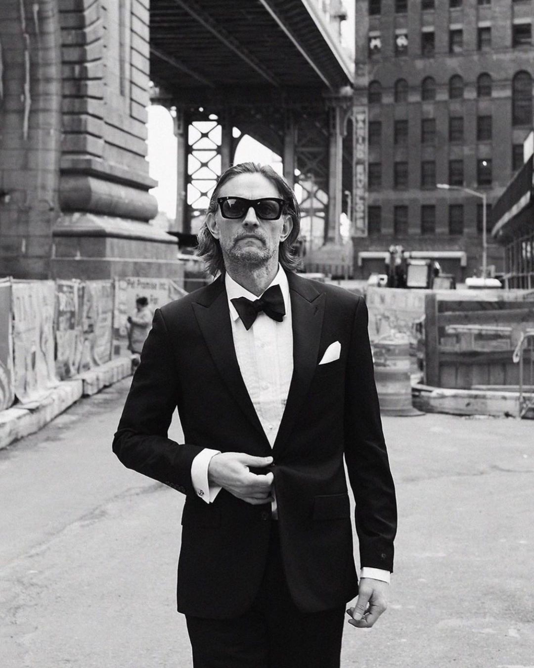 Black and white portrait of a mature man in a tuxedo with a bow tie, posing outdoors under a bridge with buildings in the background.