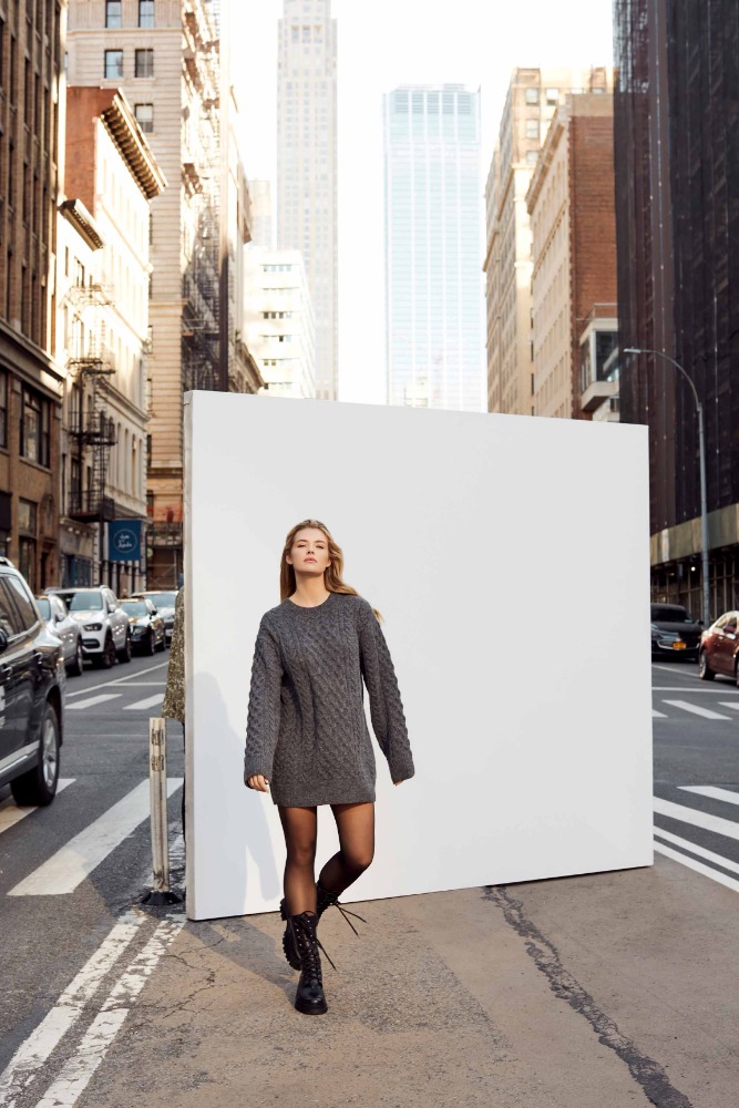 A blonde woman in a gray dress walks toward the camera in a city street with tall buildings and cars in the background.