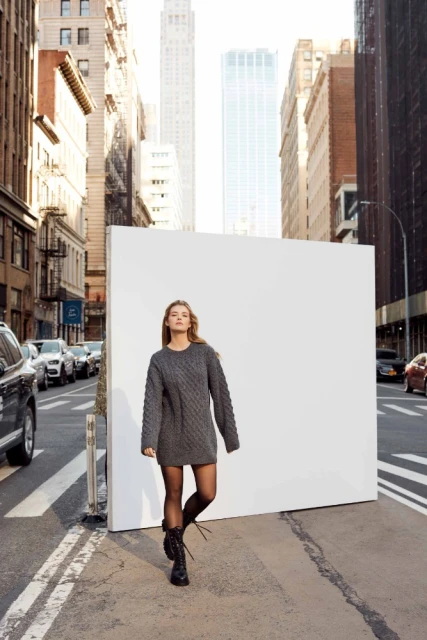A blonde woman in a gray dress walks toward the camera in a city street with tall buildings and cars in the background.