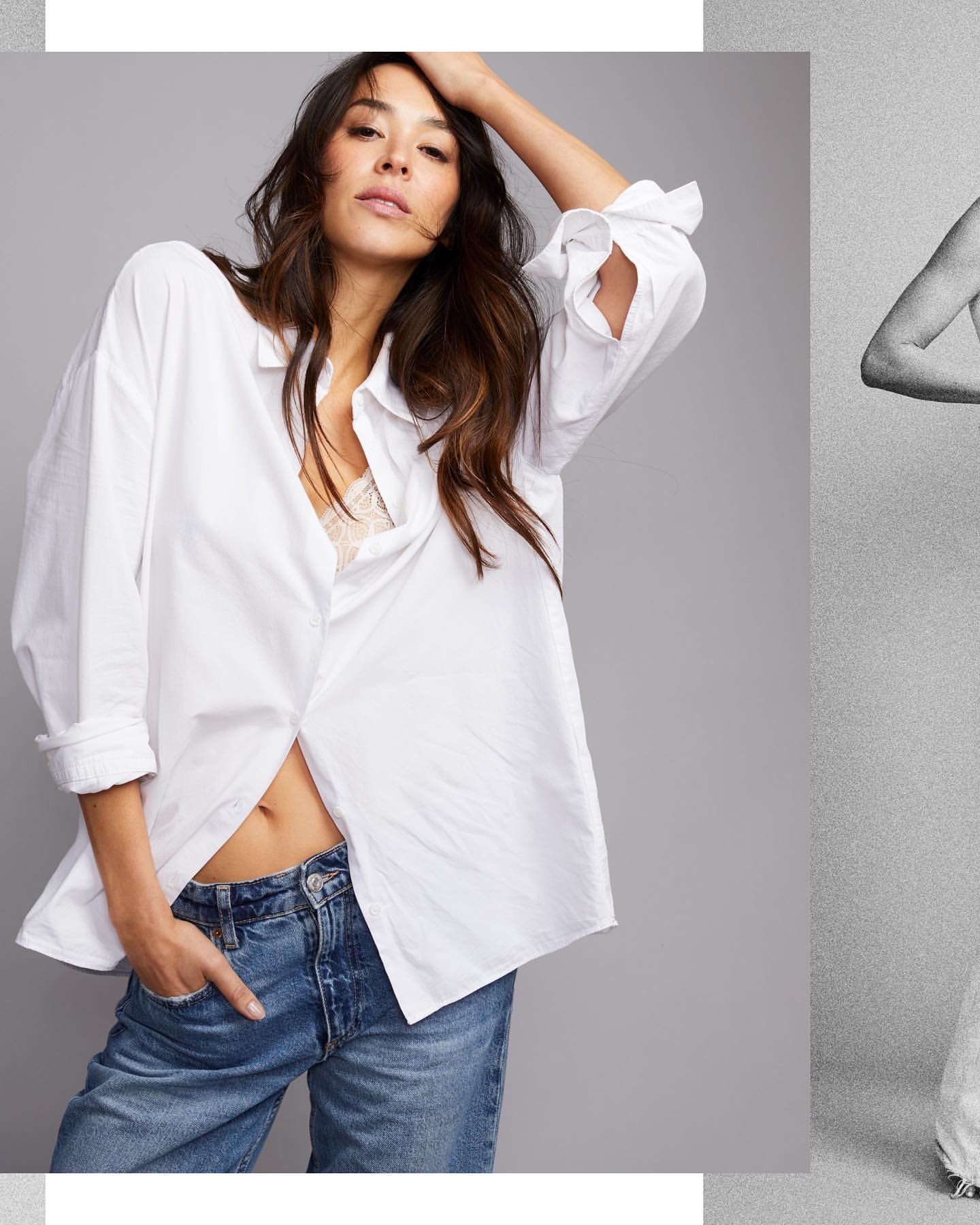 A brunette woman poses in a photo studio. She wears a white, oversized shirt and blue jeans, and looks directly at the camera.