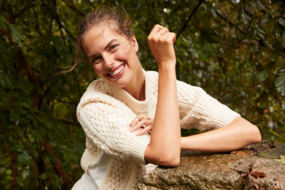 Smiling adult female model wearing a cream sweater, posing outdoors in a forest setting on a rock with a happy facial expression.