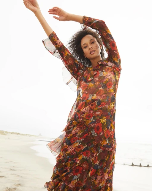 Woman poses in a long-sleeved floral maxi dress on a beach with white sand and gentle waves, showcasing fashion apparel in natural light.