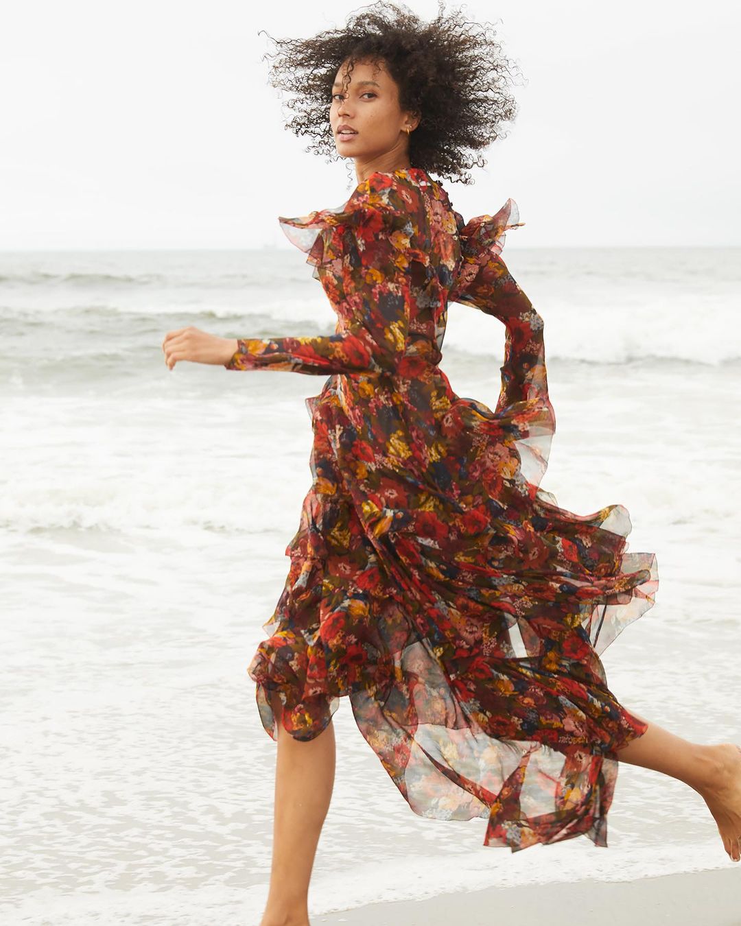 A woman with dark curly hair runs on a beach wearing a long-sleeved, floral print dress with a ruffled neckline, with ocean in background.