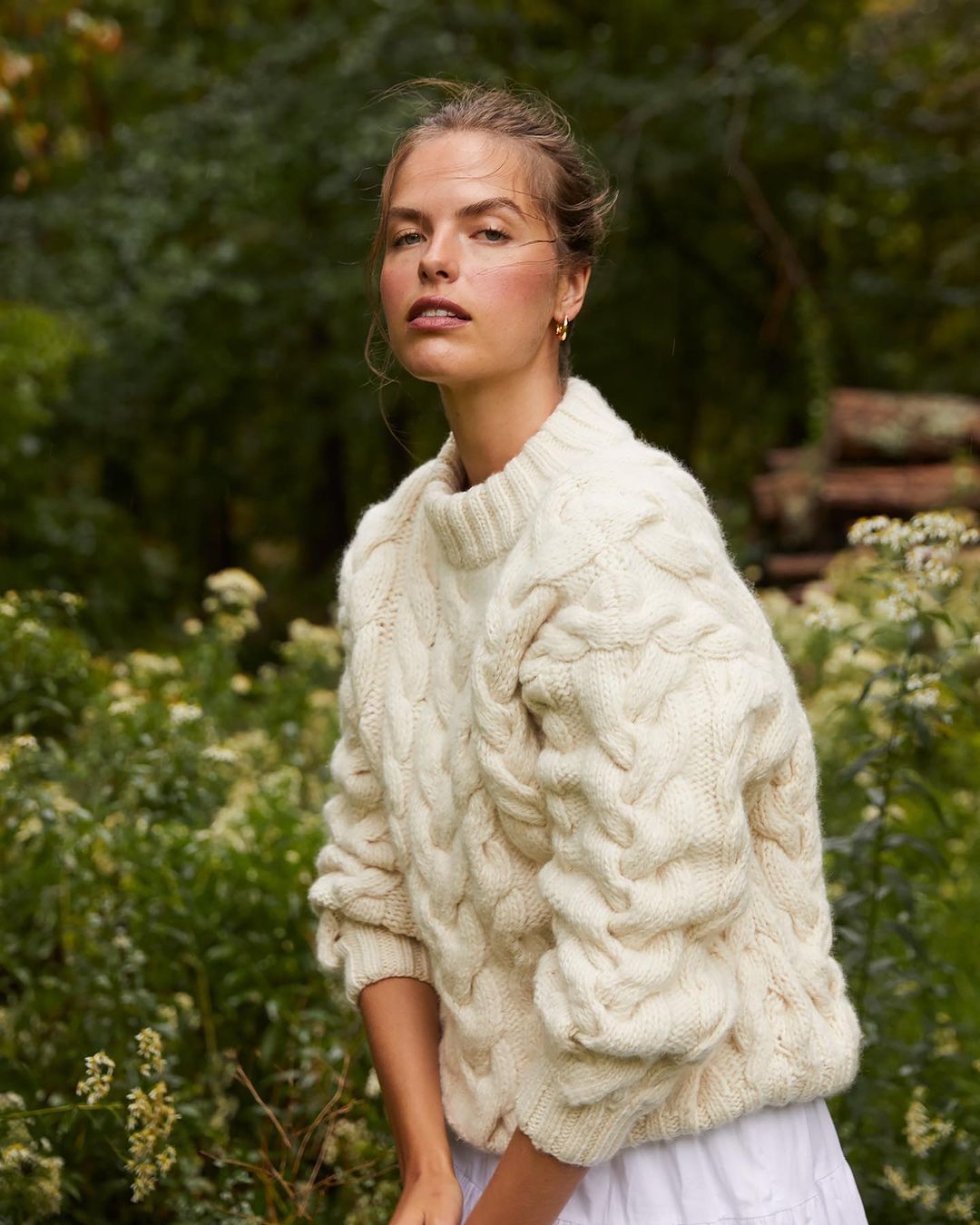 A woman stands in a field wearing an off-white cable-knit sweater and a white skirt with a background of greenery.