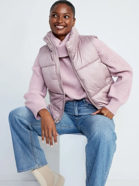 Smiling woman in a lilac puffer vest, pink sweater, and blue jeans sits in a studio against a white background.