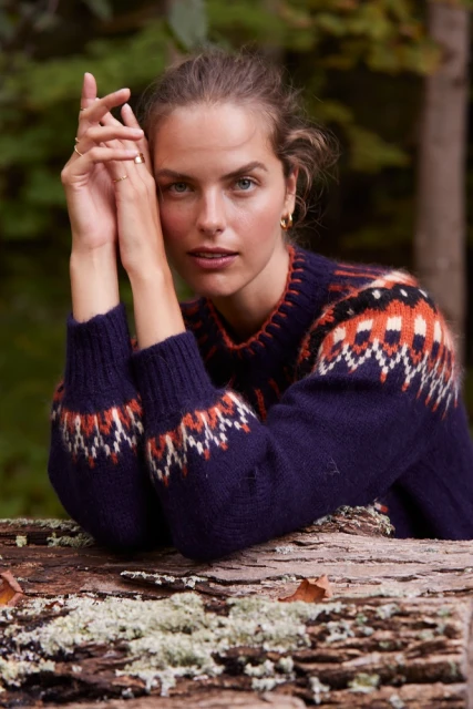 Close-up of a woman with fair skin wearing a blue sweater with a pattern leaning on a moss-covered log in a wooded outdoor setting.