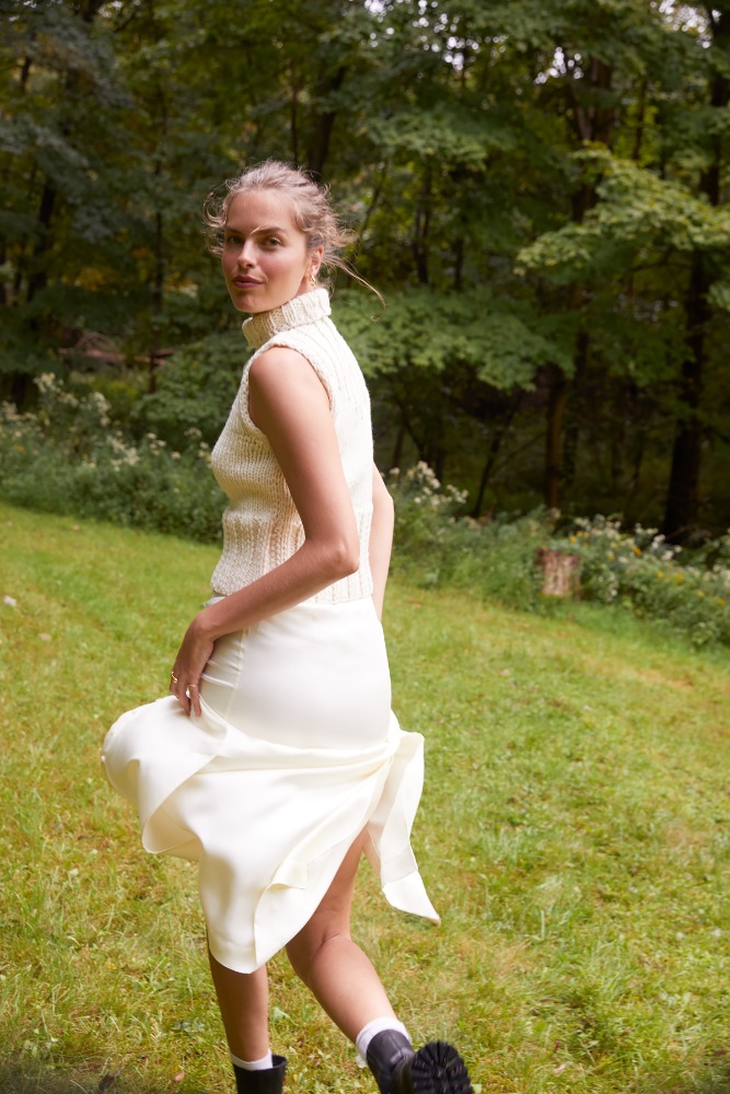 Woman in cream knit top and skirt, black boots strides across grassy clearing with trees. Soft light, fashion editorial shoot.