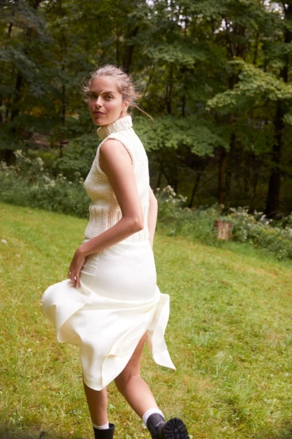 Woman in cream knit top and skirt, black boots strides across grassy clearing with trees. Soft light, fashion editorial shoot.
