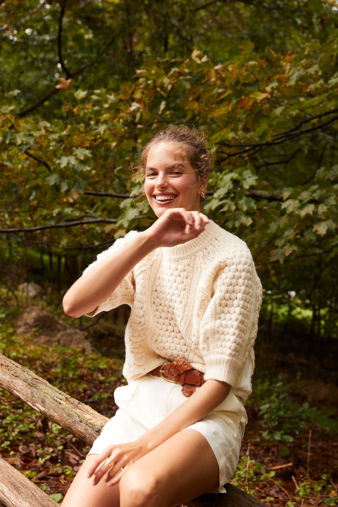 A smiling adult woman in a cream sweater and white shorts sits on a fallen tree in a forest setting, with dappled lighting.