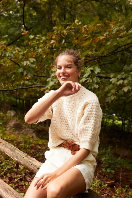 A smiling adult woman in a cream sweater and white shorts sits on a fallen tree in a forest setting, with dappled lighting.