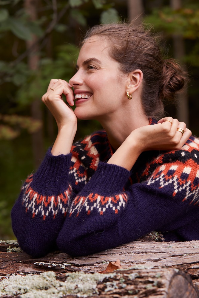 Smiling woman in navy fair isle sweater resting chin on hand outdoors with a forest background