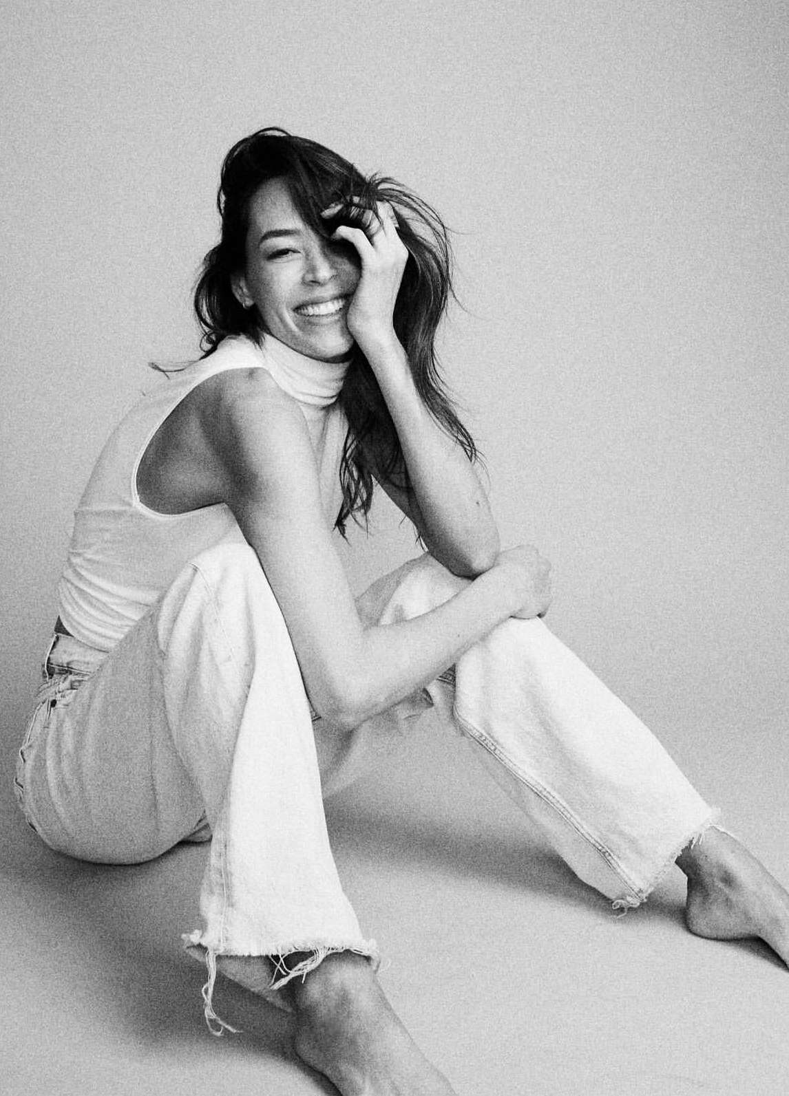 Monochrome portrait of a smiling woman sitting on the floor wearing a sleeveless turtleneck top and light-wash jeans.